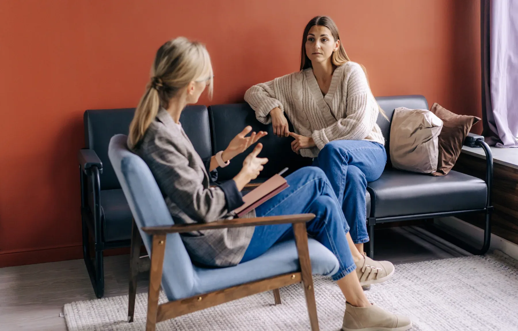 A blonde therapist in a plaid blazer and jeans sits in a light-blue wooden armchair, gesturing with both hands while holding