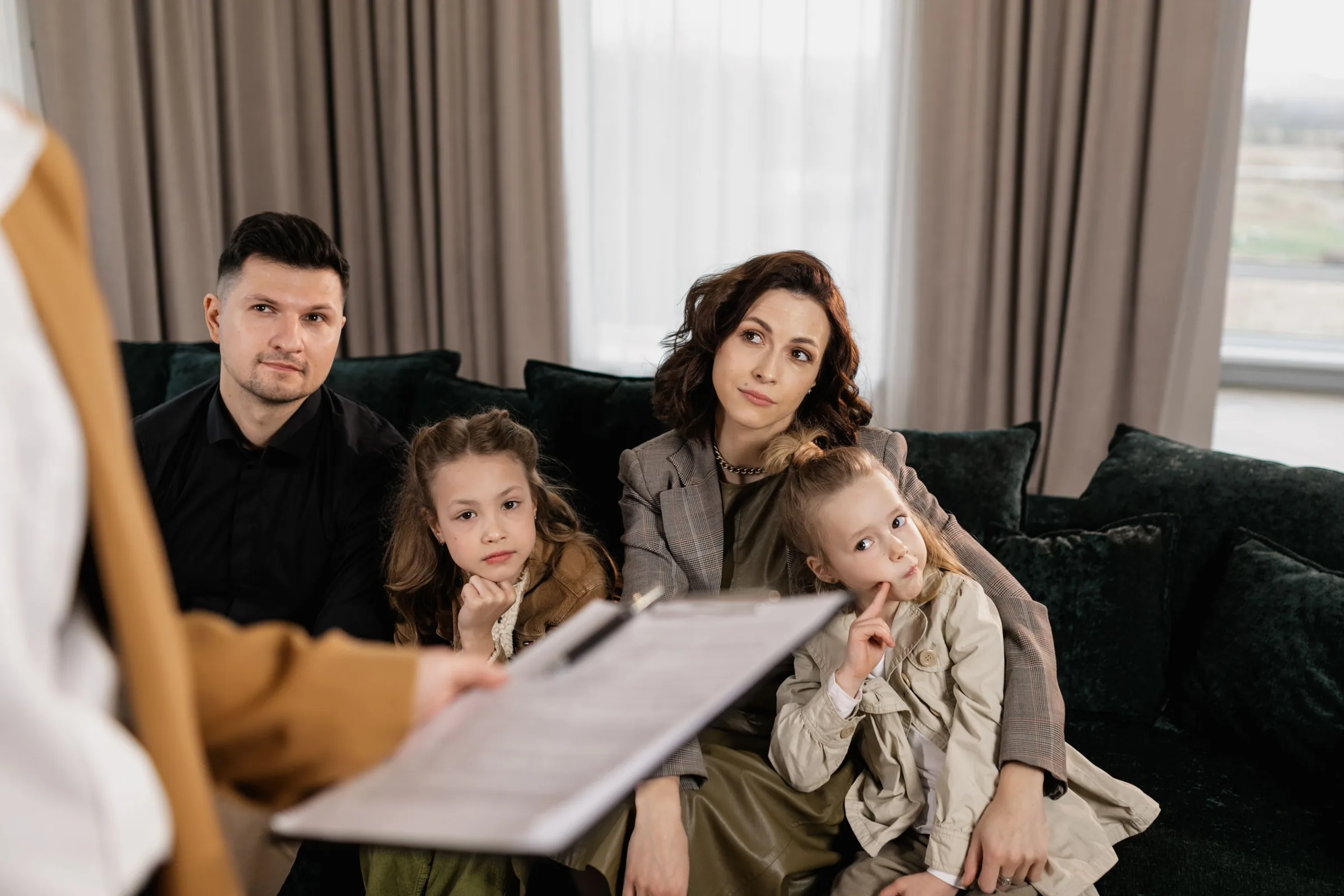Family of four sitting on a couch listening to a therapist holding a clipboard during a session.