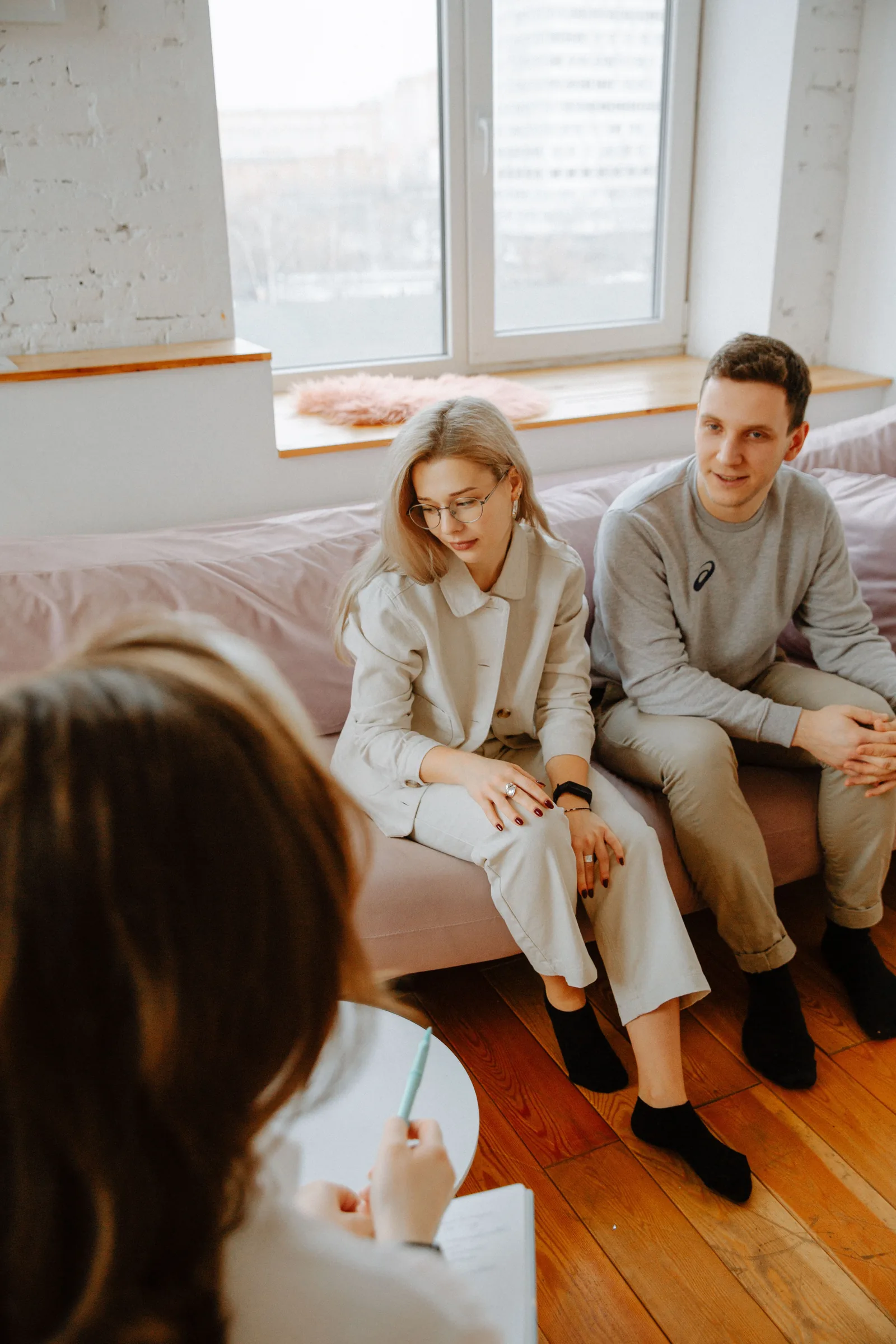A psychotherapist listens to a couple during a therapy session in a bright, modern office with large windows.