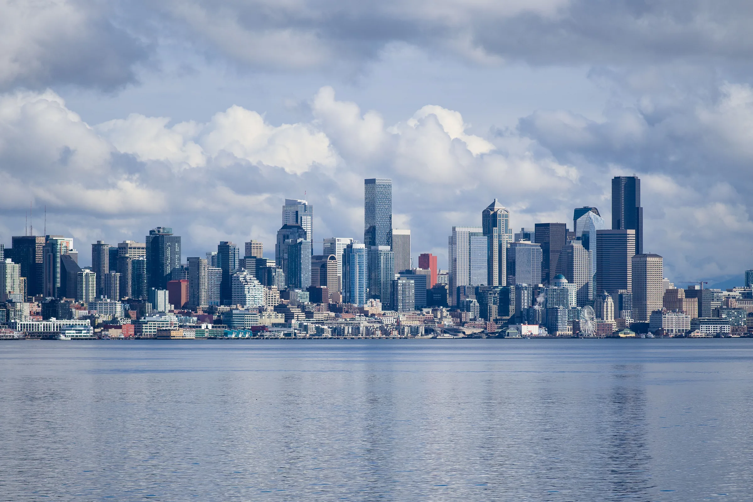 Seattle skyline with tall buildings reflected in calm blue water under cloudy sky.