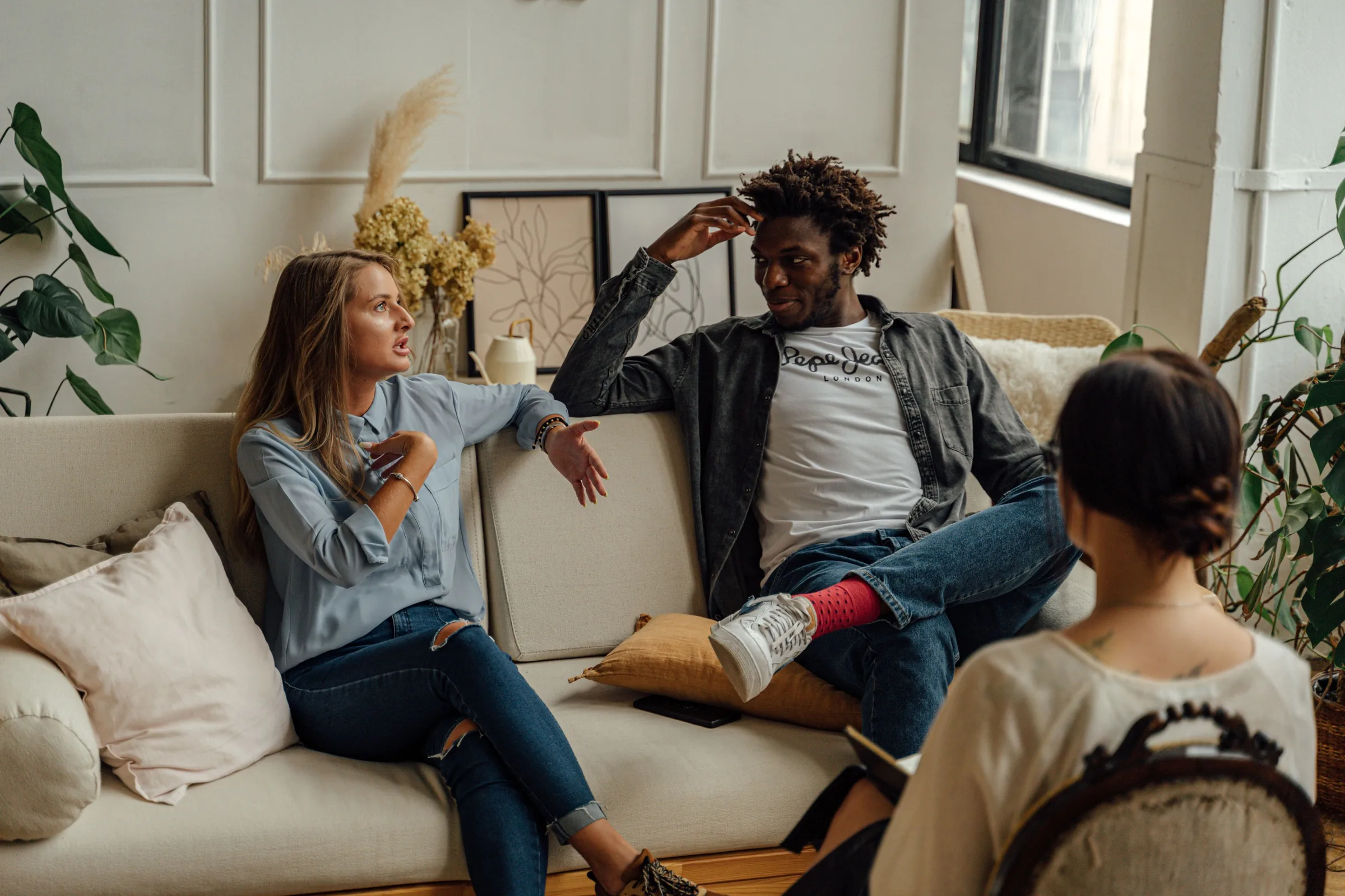 Three people sitting on a beige couch in a bright, plant-filled therapy office having a conversation.
