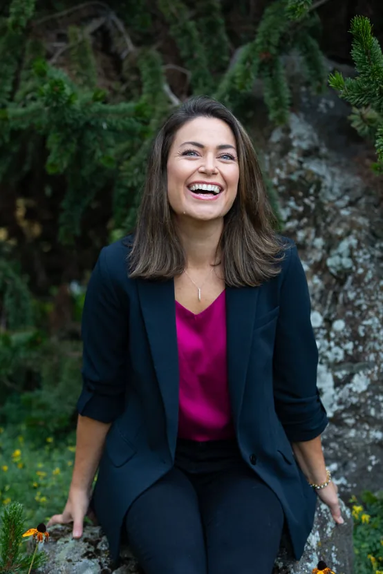 Woman in professional navy blazer and magenta top smiling outdoors against ivy-covered stone wall