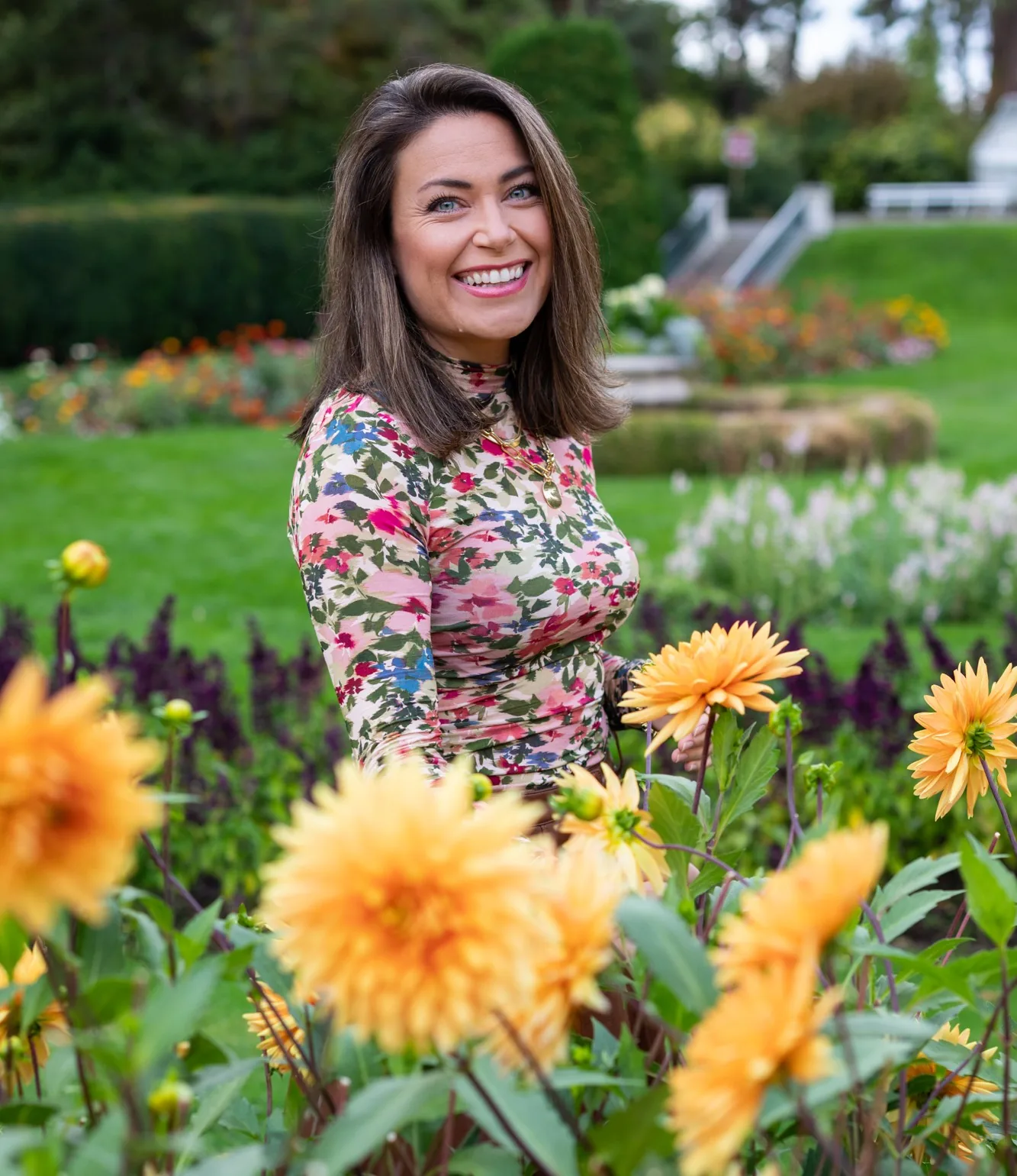 Woman smiling in a colorful flower garden with yellow gerberas and purple flowers.